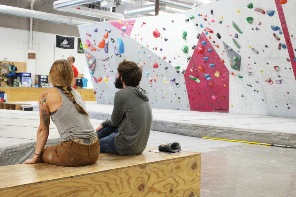 Woman in tank top and braid sits next to a man in a beard and a sweatshirt as they look at a climbing problem.